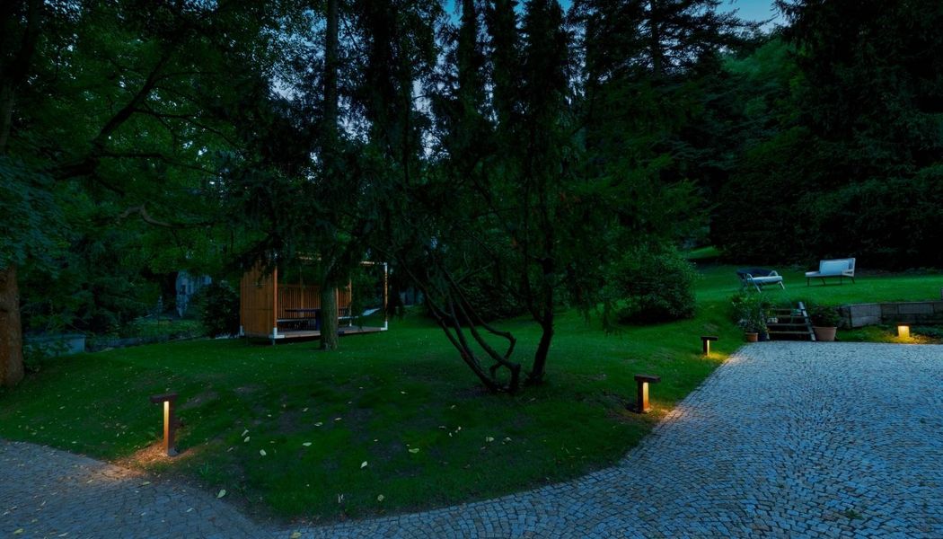 Abendansicht eines Gartens mit Bäumen, einem Kopfsteinpflasterweg und RUSTY® PATHLIGHT, die den Bereich beleuchten.