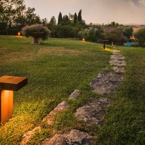 Ein Steinweg, der in der Dämmerung von RUSTY® PATHLIGHT auf einer Rasenfläche beleuchtet wird, umgeben von Bäumen und Sträuchern.