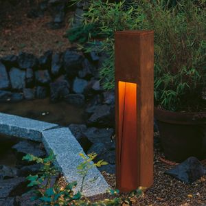 RUSTY® SLOT strahlt neben einem kleinen Teich und üppigem Grün ein warmes Licht aus und schafft so eine ruhige Gartenatmosphäre.