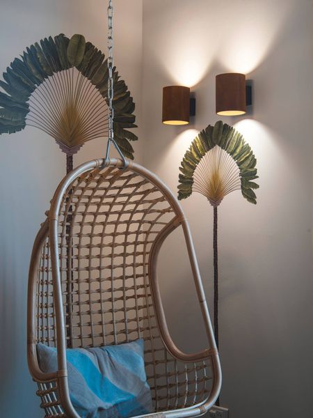 Ein hängender Korbsessel mit einem blauen Kissen, umgeben von dekorativen, an der Wand befestigten Palmwedeln und beleuchtet von zwei RUSTY® UP/DOWN.