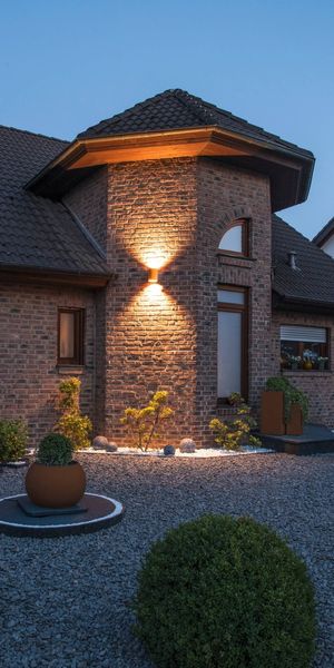 Ein Backsteinhaus mit von RUSTY® UP/DOWN beleuchtetem Eingang in der Abenddämmerung, umgeben von einem Kiesgarten mit gepflegten Sträuchern und Pflanzkübeln.