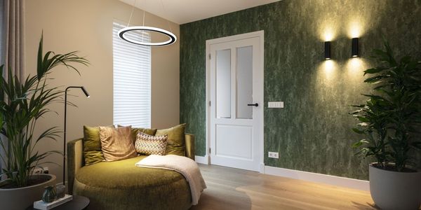 Gemütliches Zimmer mit einer grünen Akzentwand, einem runden grünen Sofa, Pflanzen, einem Beistelltisch und ONE FLAT Pendelleuchte. Heller Holzboden und weiße Tür.