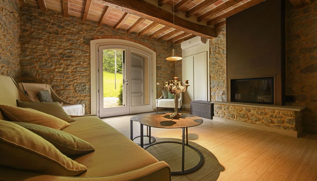 Ein rustikales Wohnzimmer mit Steinwänden, Holzbalken, einem Kamin und gewölbten Glastüren. Zur Einrichtung gehören ein Sofa und ein runder Couchtisch, über dem eine LALU® hängt.