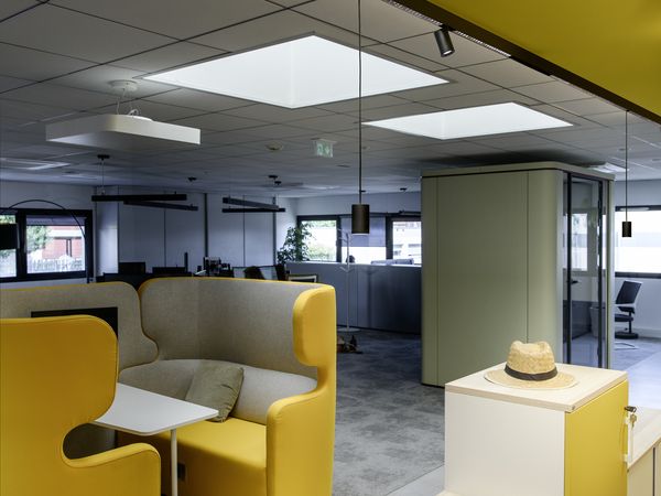 Ein modernes Büro mit gelben Sitzbereichen, weißen Spinden und einem Oberlicht. Im Hintergrund sind ein grauer Teppich und Glaswände zu sehen. 48-V-Schienenbeleuchtung mit Strahlern und Pendelleuchten prägt den Raum.