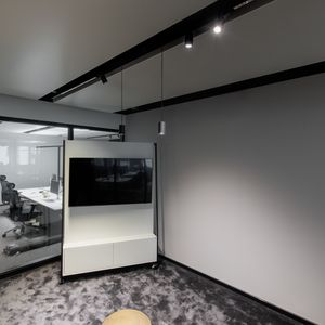 Moderner Büroraum mit grauem Teppich und minimalistischem Mobiliar, ausgestattet mit einem an der Wand montierten Fernseher und einem Blick in einen Konferenzraum mit Stühlen, beleuchtet durch 48-V-Schienenstrahler und Pendelleuchten.