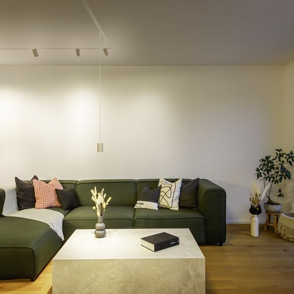 Modernes Wohnzimmer mit einem grauen Ecksofa, weißen Wänden, einem an der Wand montierten Fernseher und einem großen Fenster. Auf drei runden Tischen steht eine Vase, beleuchtet von eleganten 48-V-Schienenleuchten.