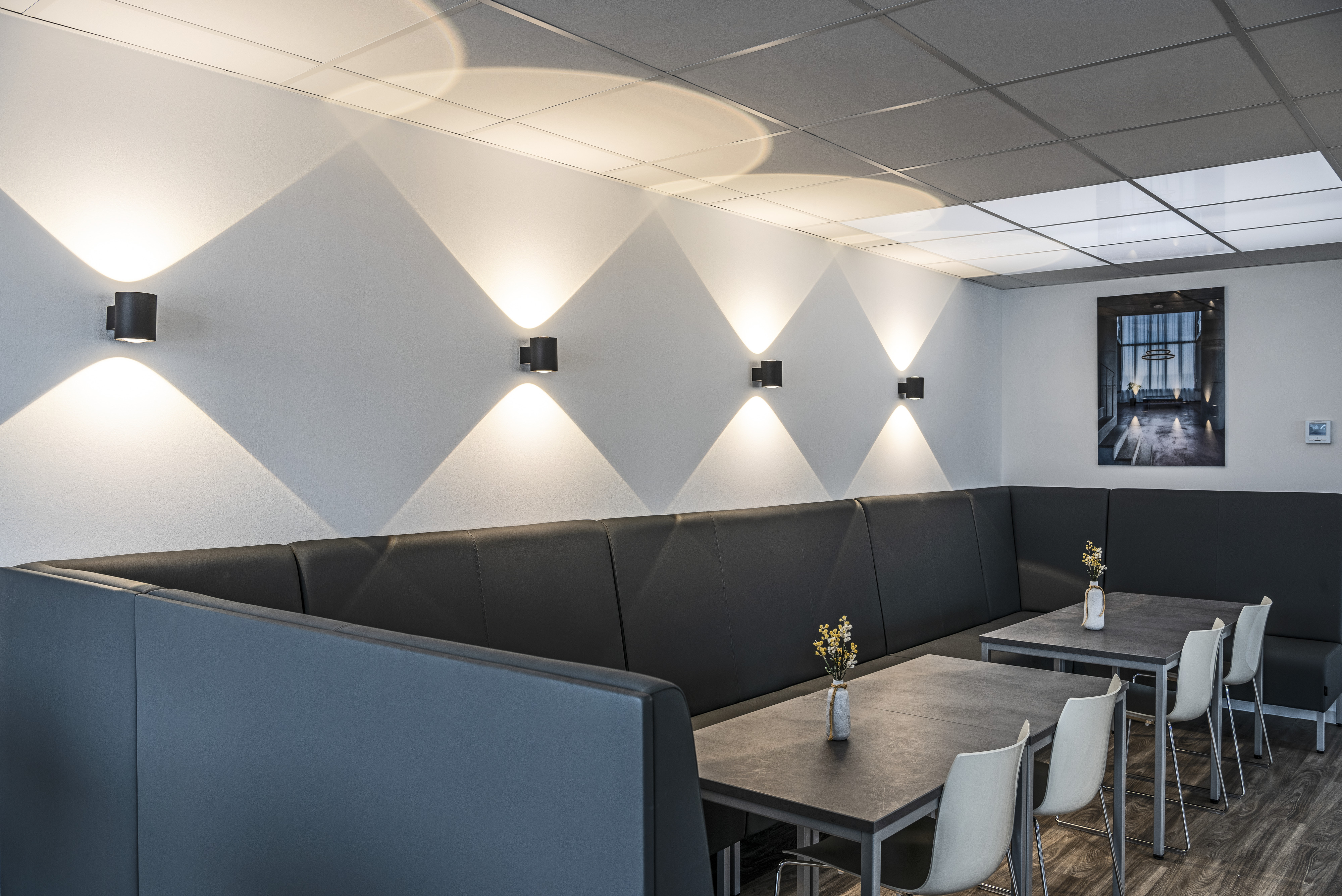Modern cafe interior with gray booths, white chairs, and tables. Wall lights cast patterns on the wall. A framed picture hangs on the wall.