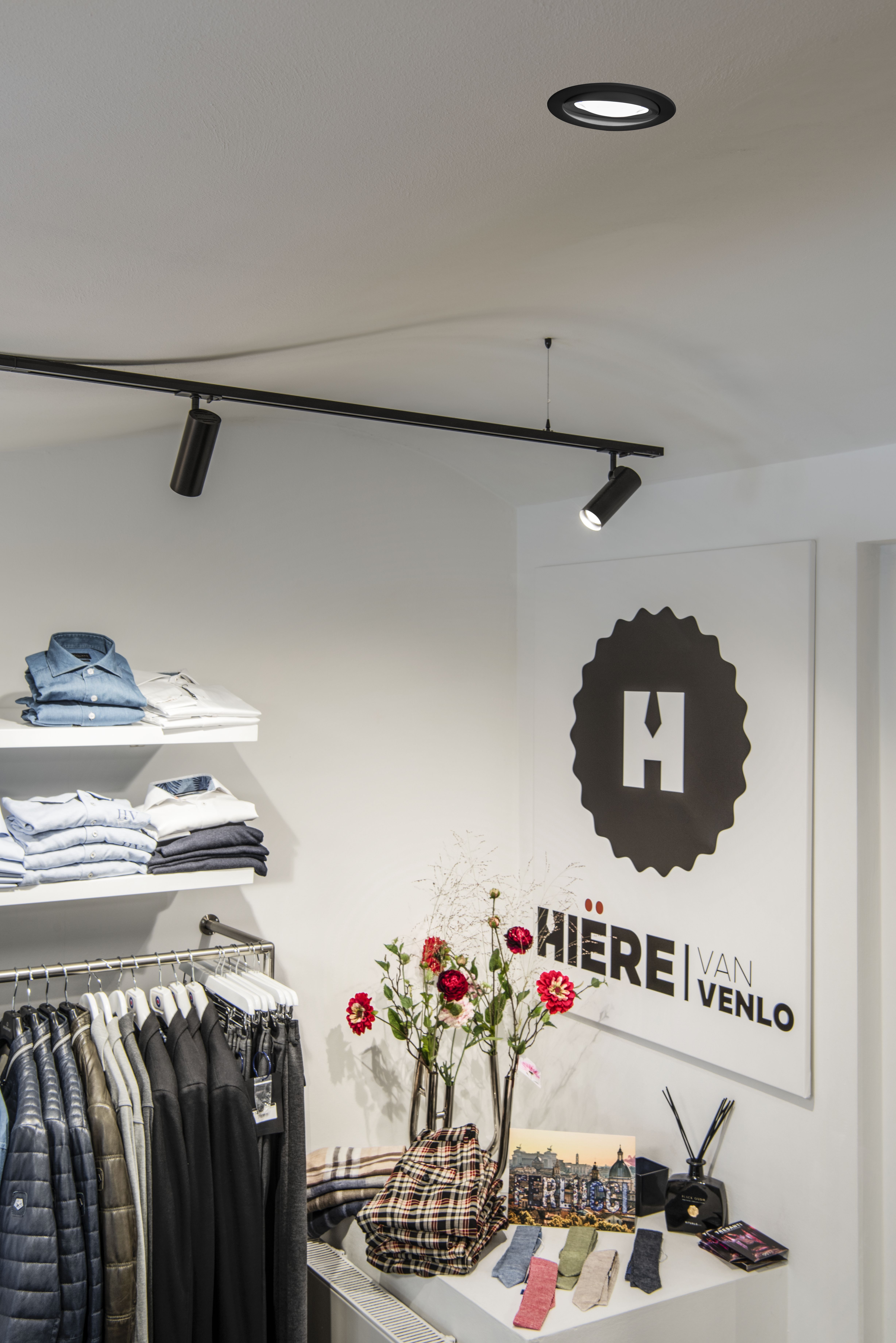 Innenraum eines Bekleidungsgeschäfts mit Jacken und Hemden in Regalen, einem Ausstellungs-Tisch mit Blumen, Socken und einem Schild „Hiere van Venlo” an der Wand. Beleuchtet mit OCULUS Einbauleuchten als Grundbeleuchtung.