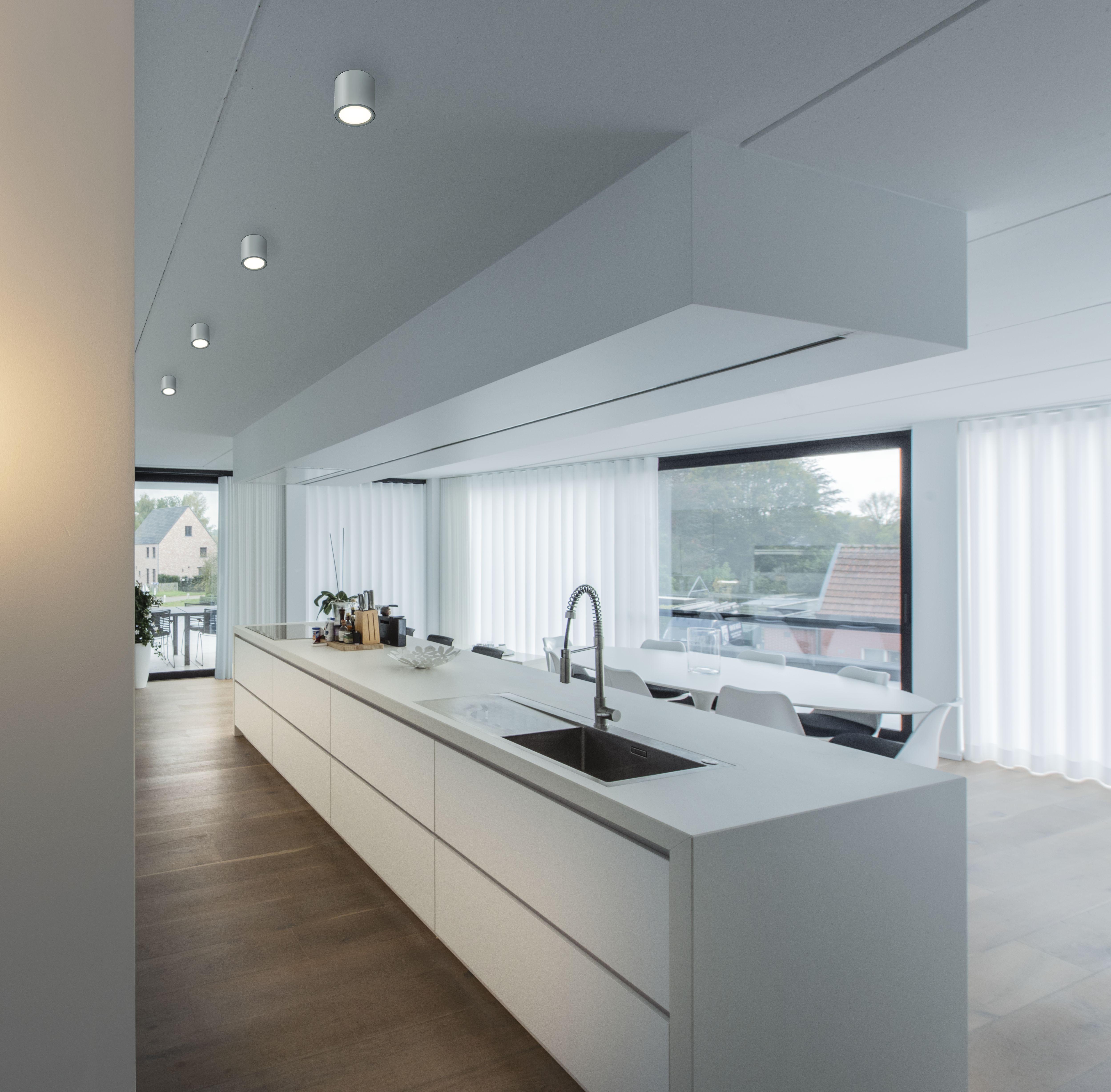 Modern kitchen with a long white island, built-in sink, wooden floor, and large windows with sheer curtains, offering a bright, open space.