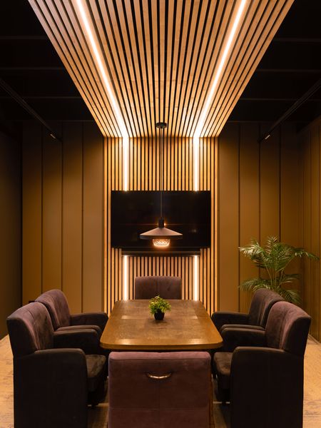 Modern conference room with a wooden table and LALU® pendant set, cushioned chairs, a wall-mounted TV, warm lighting, vertical wood paneling, and a potted plant.