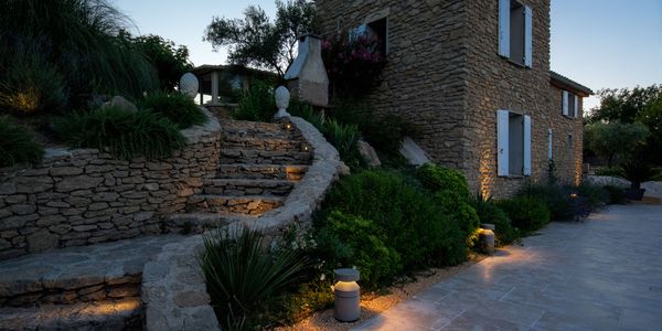 Stone house at dusk with illuminated steps and CONCRETE path light, surrounded by lush greenery and a clear evening sky.