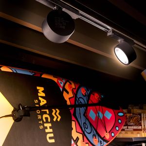 Colorful surfboard with "MARCHU SPORTS" logo displayed under KALU 2.0 spotlights in a store, with shelves of products in the background.