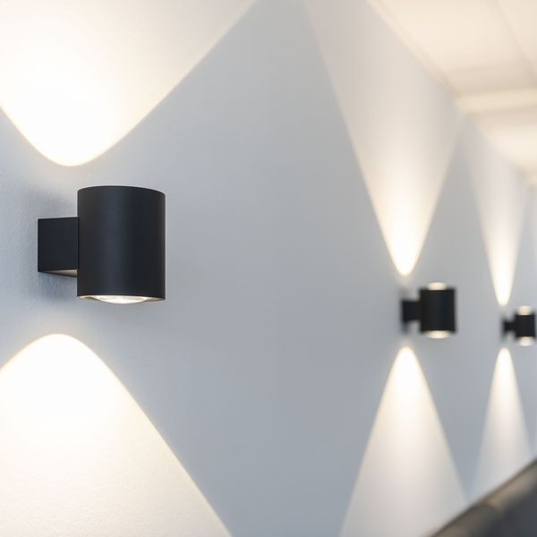 Wall-mounted black cylindrical lights casting intersecting patterns on a white wall, creating a symmetrical and modern design.