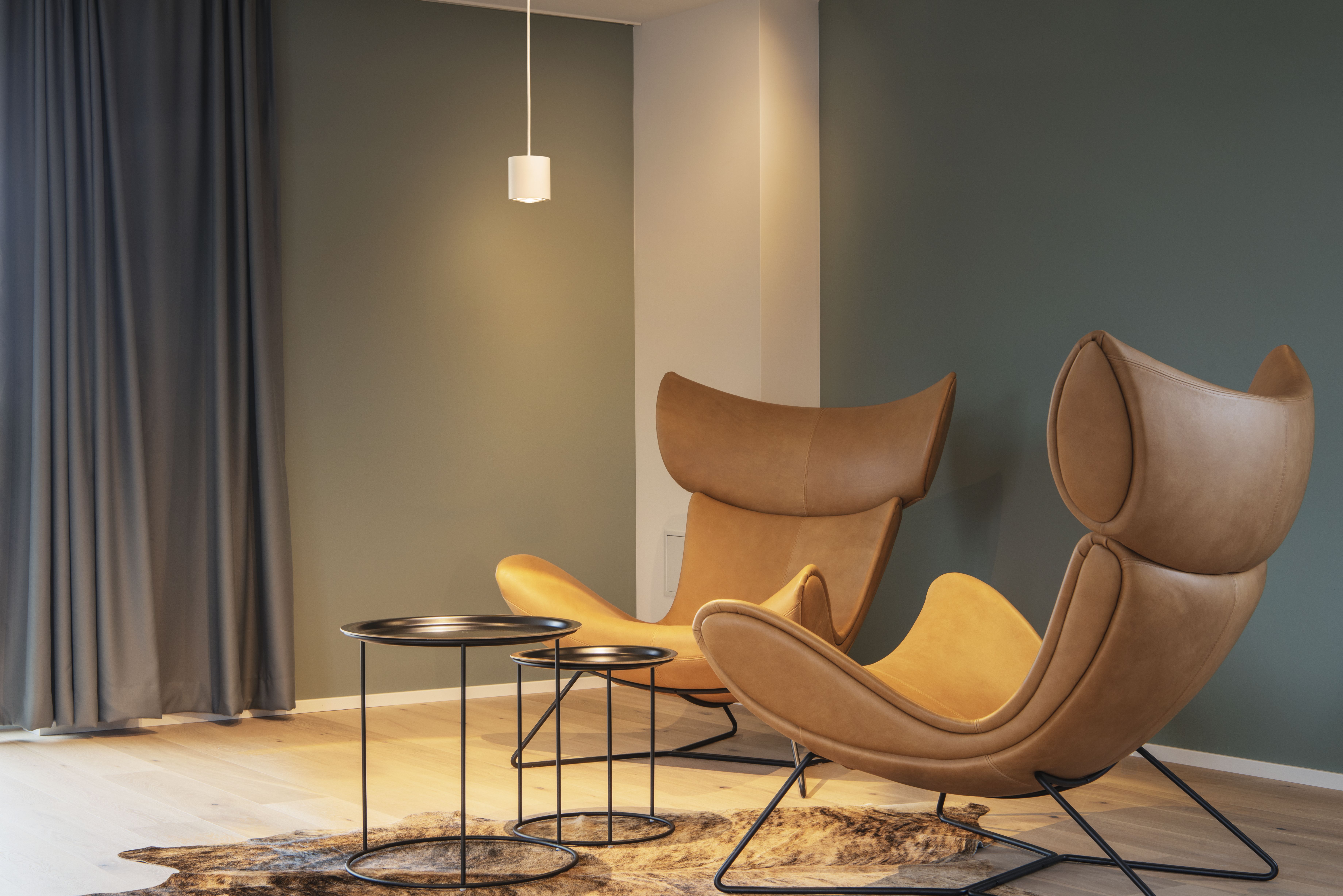 Modernes Zimmer mit zwei braunen, geschwungenen Sesseln, drei runden schwarzen Tischen, der OCULUS Pendelleuchte über dem Tisch und dunklen Vorhängen an einer grünen Wand.