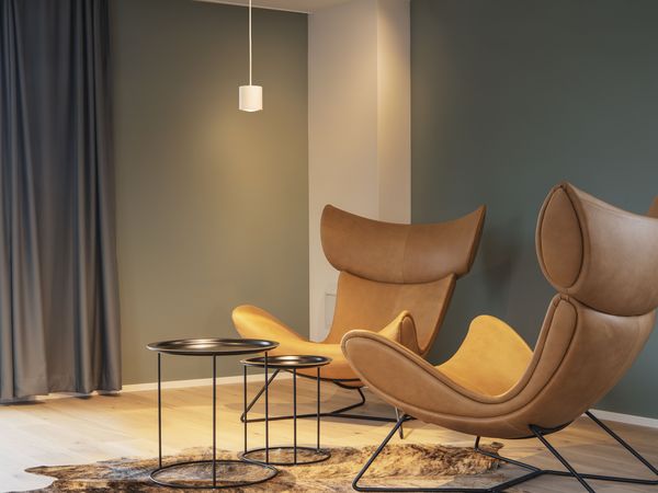 Modern room with two brown, curved armchairs, three round black tables, a hanging light, and dark curtains on a green wall.