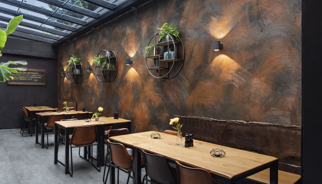 Modern cafe interior with wooden tables, leather chairs, and wall-mounted plant decor. Warm lighting and a glass ceiling create a cozy atmosphere.