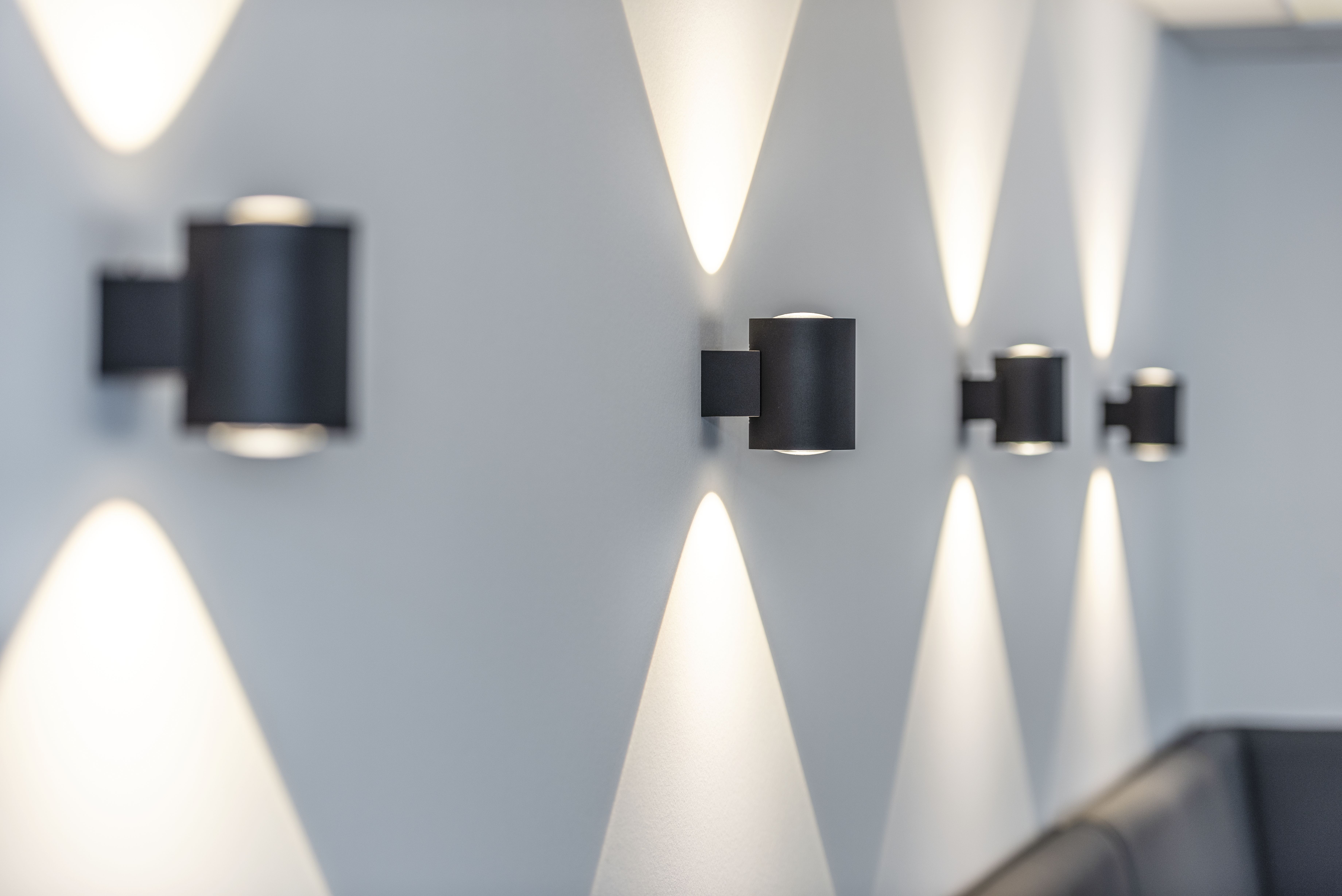 Wall with modern black cylindrical sconces casting light upwards and downwards, creating a diamond pattern on the light gray surface.
