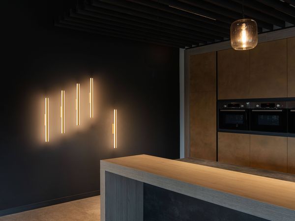 Modern kitchen with dark walls, ONE LINEAR as wall lights, wooden cabinets, built-in ovens, and a minimalist wooden island under a PANTILO pendant light.