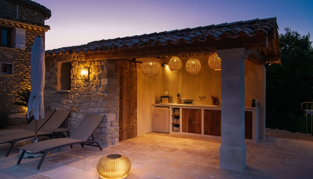 Outdoor stone patio at dusk with lounge chairs, a rustic kitchenette, hanging wicker lights, and a glowing ADEGAN on the tiled floor.