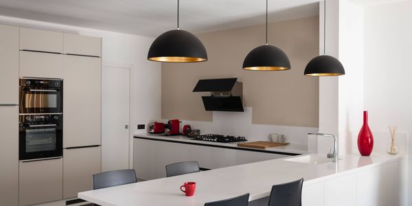 Modern kitchen with white cabinets, FORCHINI pendant lights, a long white island, and black chairs. Red accents include a vase and a mug.