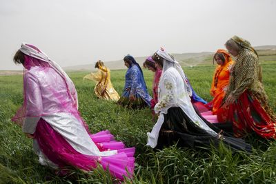 Women in vibrant traditional clothing walk through a green field, with hills in the background under a cloudy sky.