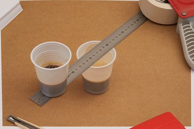 Two plastic cups of coffee on a brown surface, with a metal ruler balanced between them. Nearby are a cutter, tape, and a red stapler.