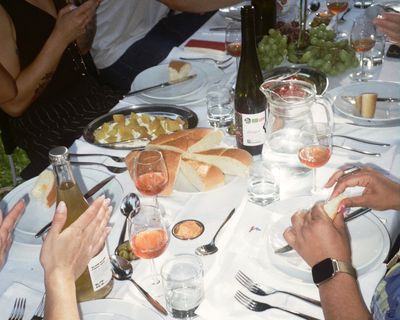 A table set for a meal with people sitting, featuring bread, cheese, grapes, wine bottles, and glasses of rosé wine. Hands are reaching for bread.