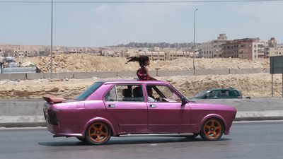 A pink car with orange wheels drives along a road; a child stands through the sunroof with hair blowing in the wind. Urban buildings are visible in the background.
