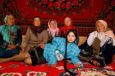 Six women, five seated wearing headscarves and one lying in front in a blue plaid outfit, pose on a vibrant red floral carpet.