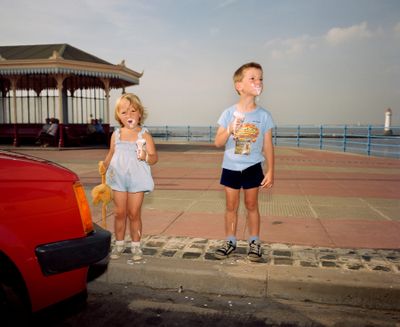 Two children with ice cream on their faces stand near a red car holding stuffed toys by the seaside, a lighthouse in the background.