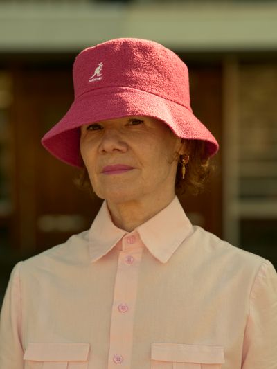 Woman in a pink shirt and matching bucket hat stands outdoors, with a neutral expression and blurred background.