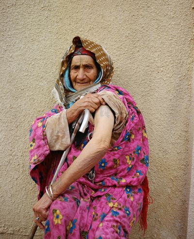 Elderly person in a vibrant pink floral dress sits with a walking cane, wrapped in a patterned headscarf, against a beige textured wall.