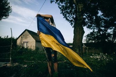 Person holding a large Ukrainian flag in a garden, with trees and a house in the background under a clear sky.