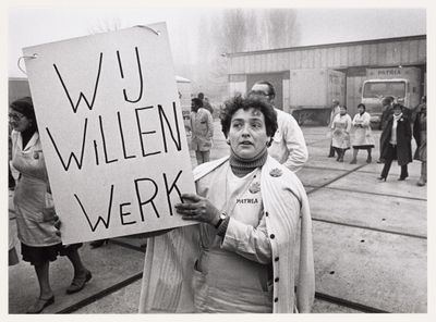 A woman holds a sign reading "Wij Willen Werk" in Dutch, meaning "We Want Work," during a protest.