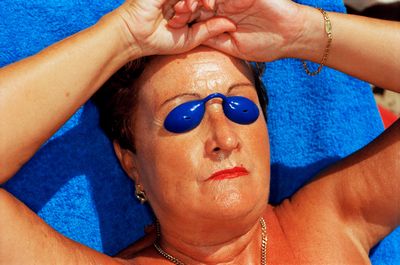 Woman sunbathing on a blue towel, wearing blue tanning goggles, with arms above her head and gold jewelry.