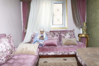 A person sitting on a plush pink sofa, using a phone in a cozy room with floral decor and sheer curtains.