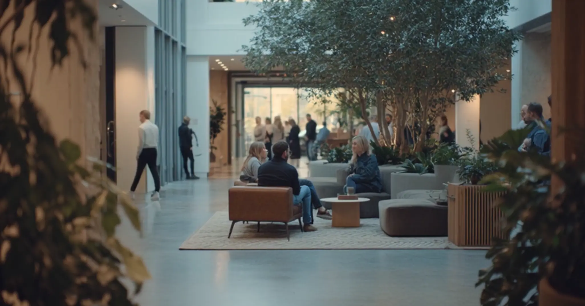 People sitting and conversing in a modern, indoor atrium with large plants, natural light, and a sleek design. Others walk by in the background.