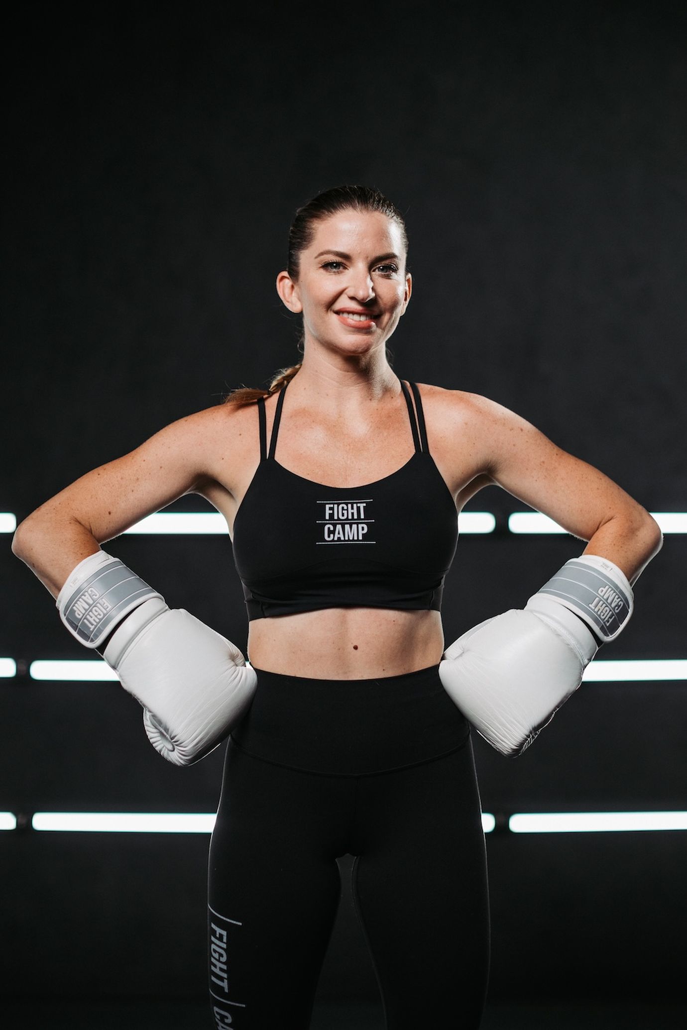 Woman in a sports bra and leggings with boxing gloves, smiling confidently against a dark background with horizontal light stripes.