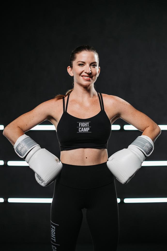 Woman in a sports bra and leggings with boxing gloves, smiling confidently against a dark background with horizontal light stripes.