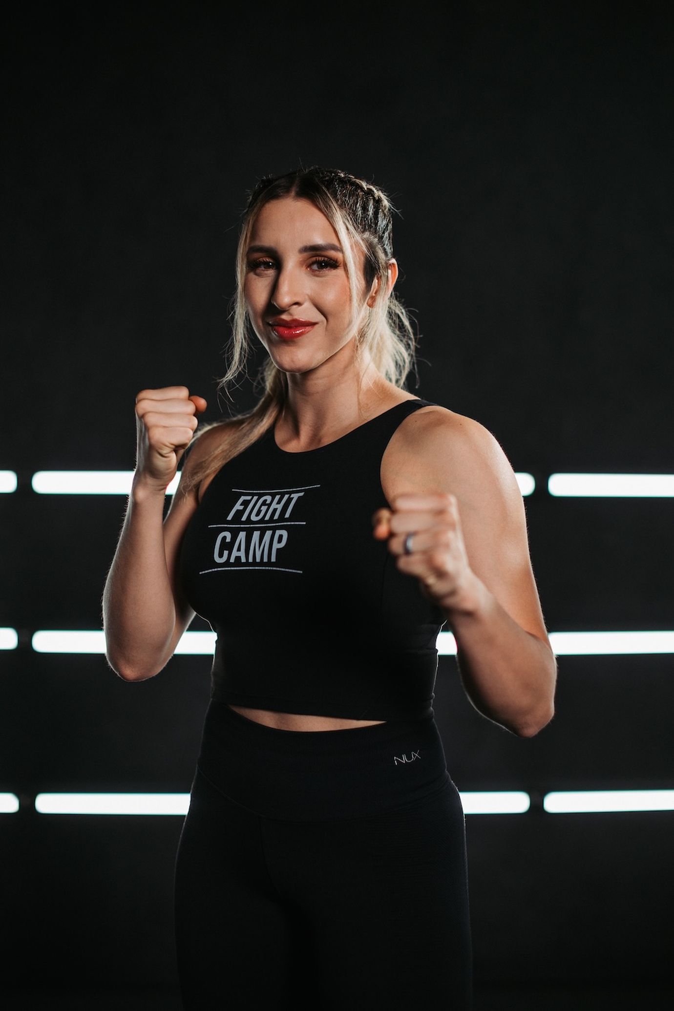 Woman in a "Fight Camp" tank top, posing with fists up in a boxing stance, against a dark background with horizontal light strips.