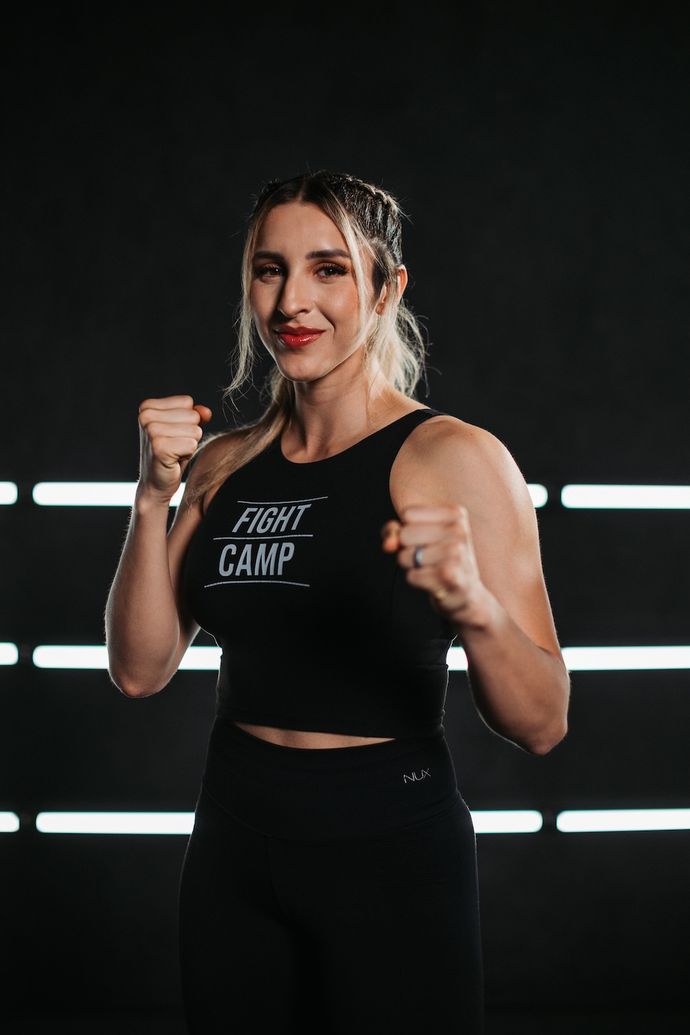 Woman in a "Fight Camp" tank top, posing with fists up in a boxing stance, against a dark background with horizontal light strips.