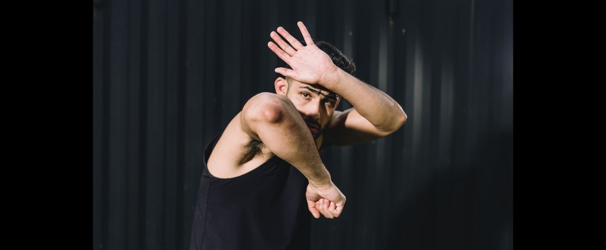A man in a black tank top poses defensively against a dark background, shielding his face with one arm and raising the other.