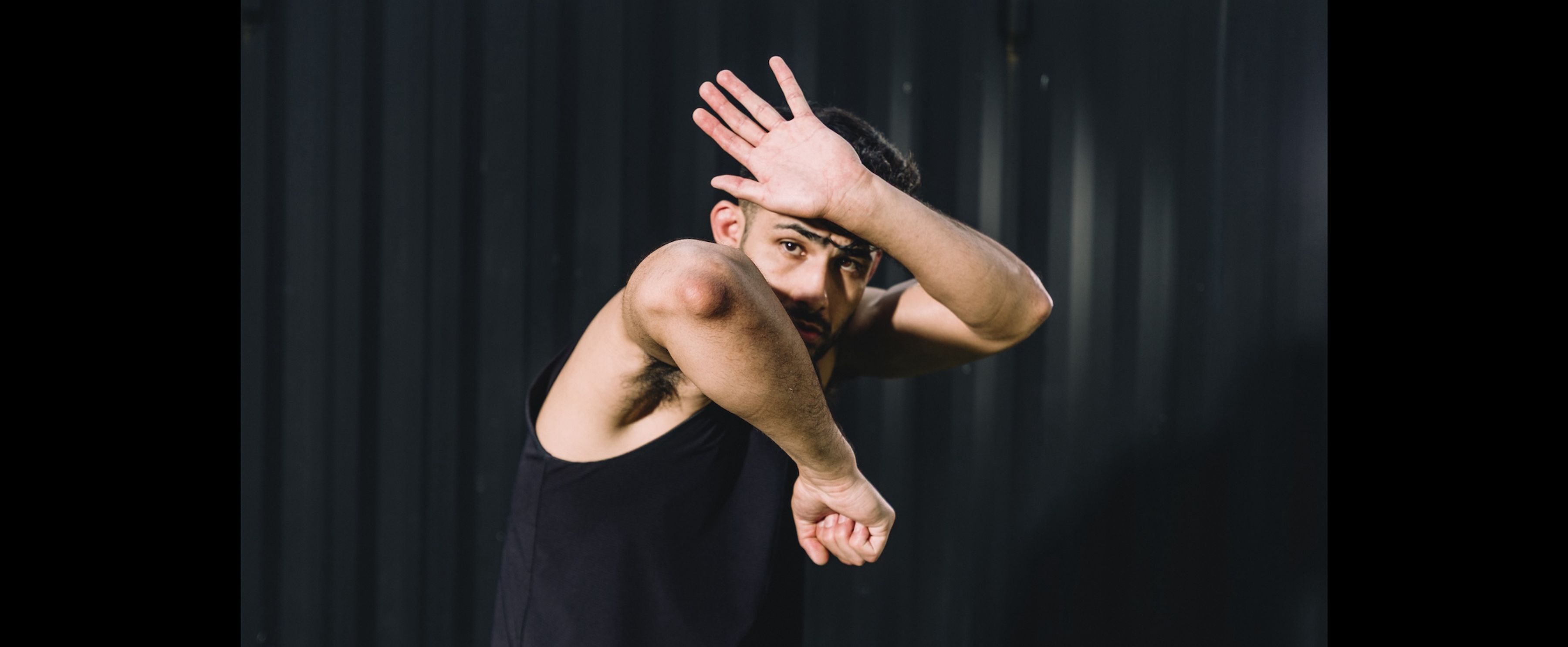 A man in a black tank top poses defensively against a dark background, shielding his face with one arm and raising the other.