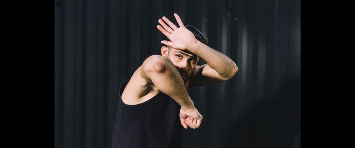 A man in a black tank top poses defensively against a dark background, shielding his face with one arm and raising the other.