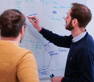 Two people collaborate at a whiteboard, discussing and writing notes and diagrams with markers.