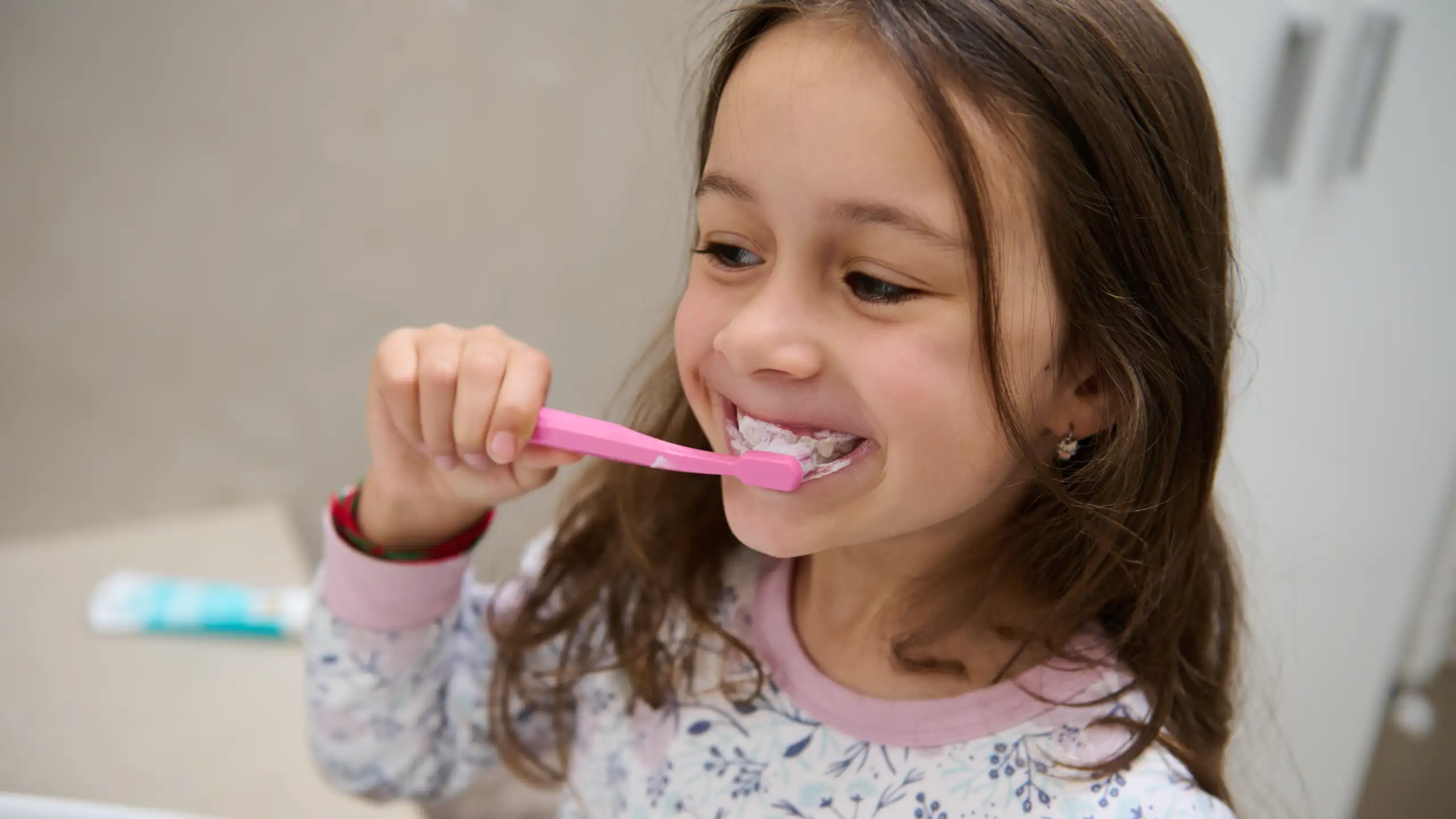 Young girl brushing her teeth with a pink toothbrush, toothpaste foam on her mouth, smiling in floral pajamas.