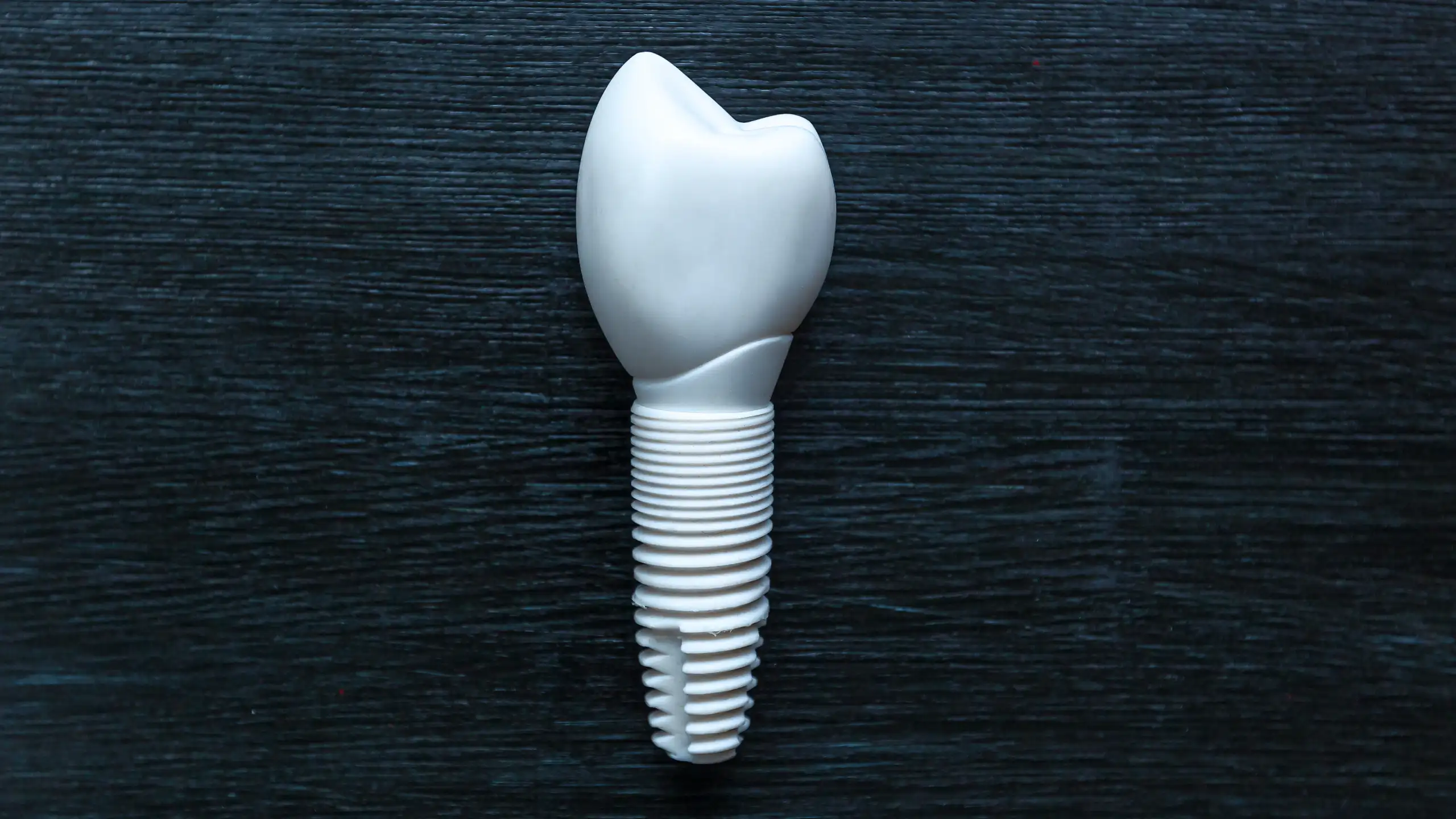 A ceramic dental implant with a tooth-shaped crown and threaded base on a dark textured surface.