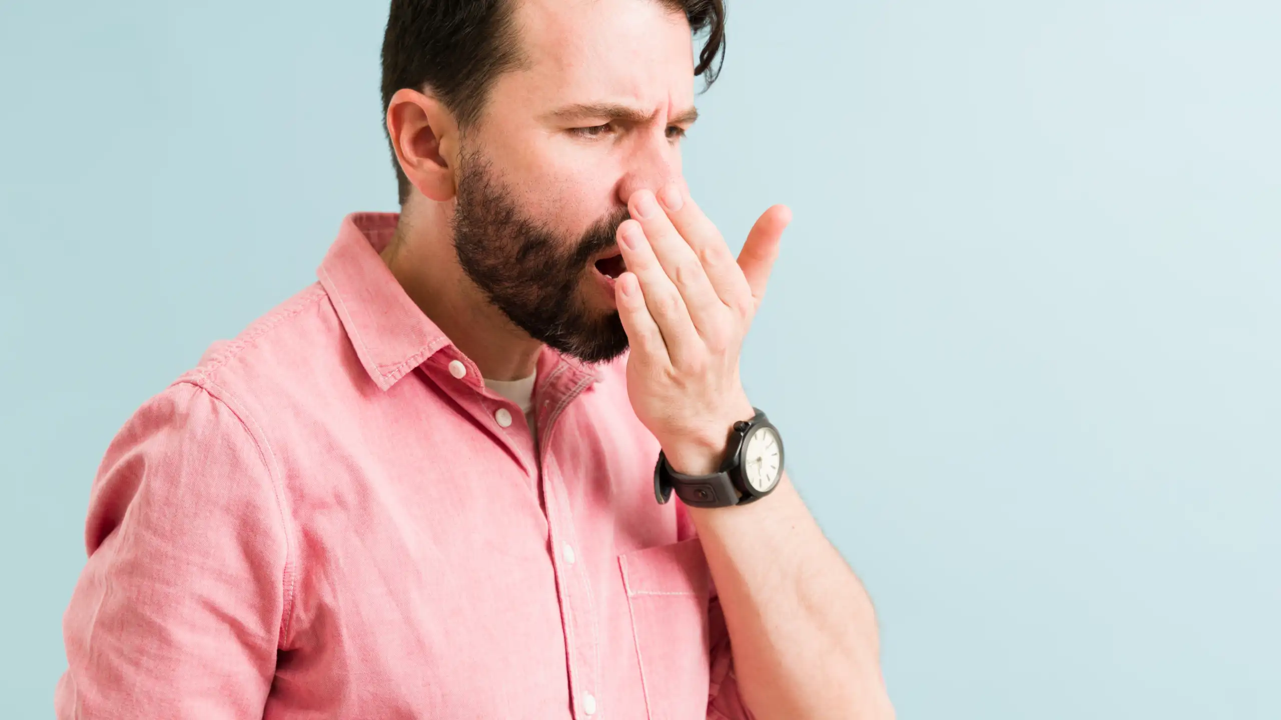 Man in a pink shirt covers his mouth with his hand, standing against a light blue background, appearing concerned or thoughtful.