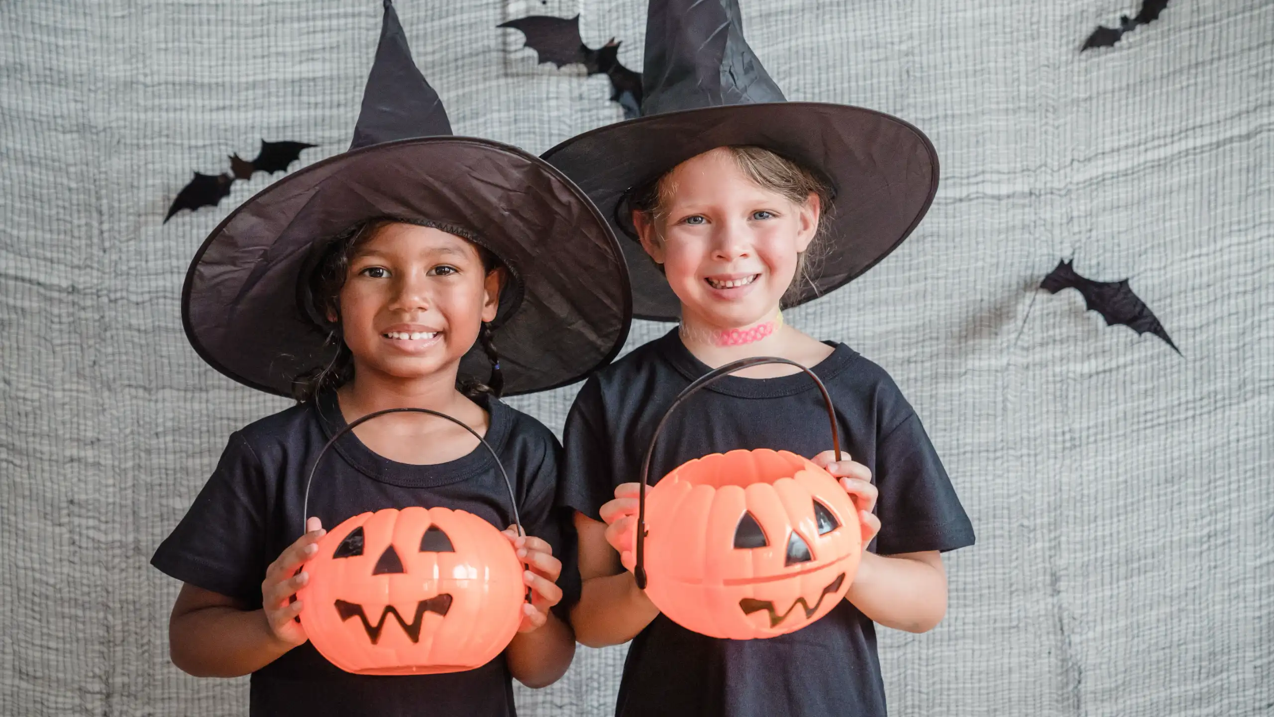 Two children dressed as witches holding orange pumpkin buckets, smiling against a backdrop with bat decorations.