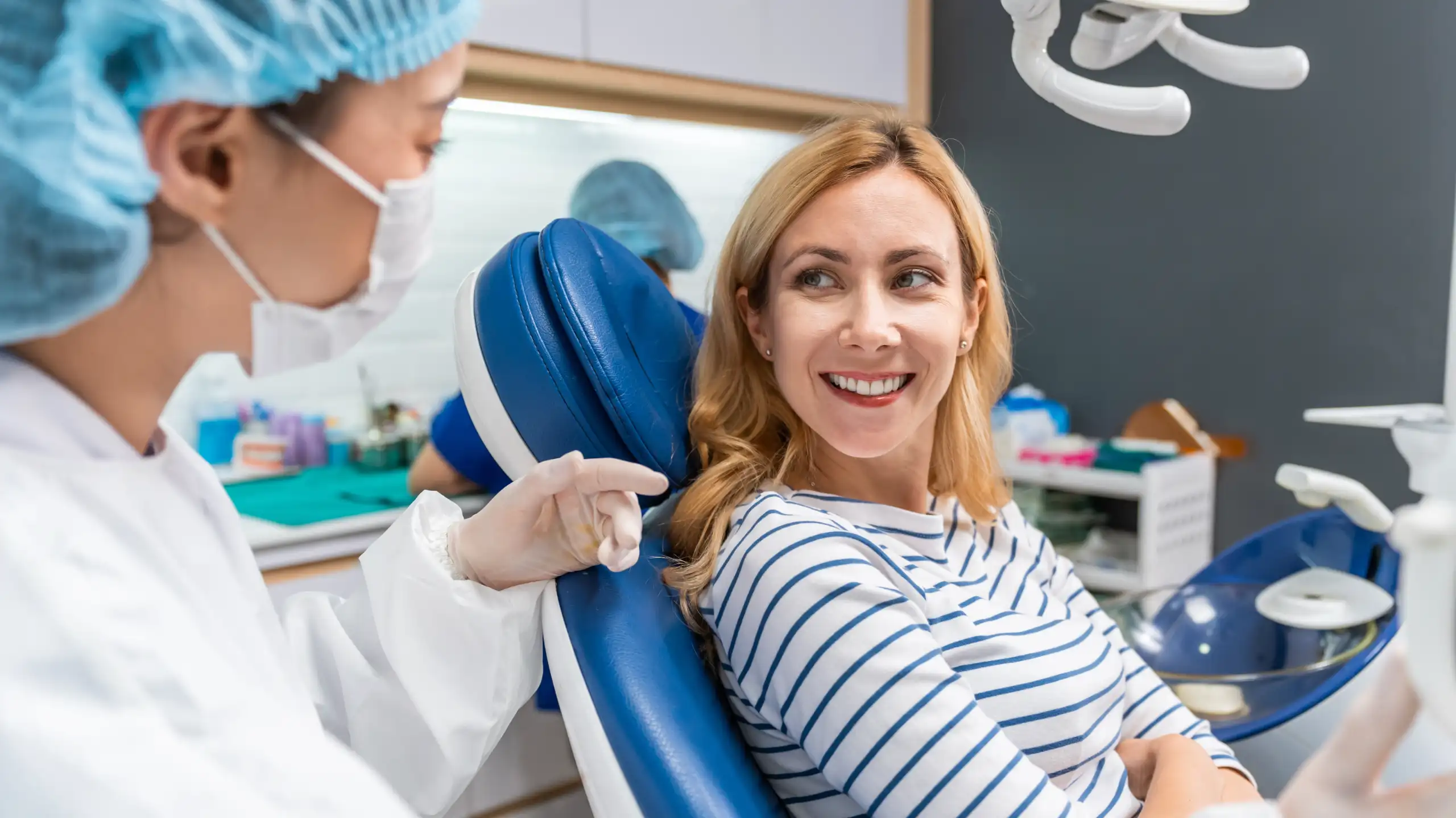 A woman in a striped shirt smiles at a masked dentist in a dental office.